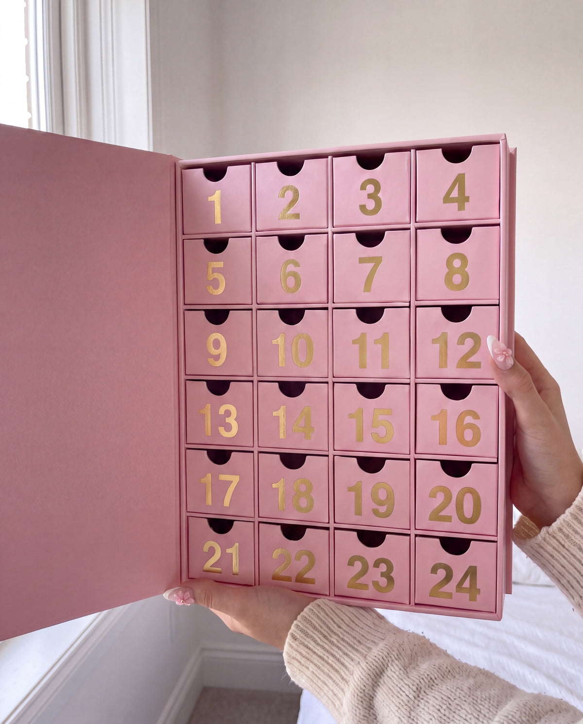 24 cases — Une main tient le calendrier de l'Avent bijoux ouvert, avec ses tiroirs numérotés.