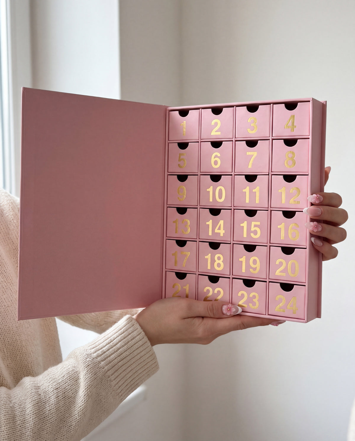24 cases — Une main tient le calendrier de l'Avent ouvert, les tiroirs dorés numérotés bien visibles.